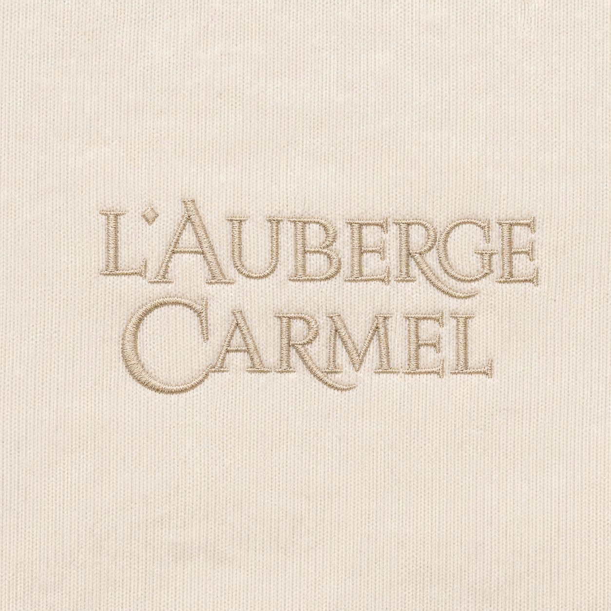 Stone-beige two-line L'Auberge Carmel wordmark embroidery on ecru jersey, macro.
